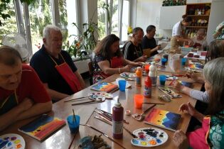 Kilka osób siedzi przy dużym stole i maluje obrazy farbami. Na stole widać palety, kubeczki z wodą i butelki z farbami. Uczestnicy noszą fartuszki ochronne i skupiają się na swoich pracach.