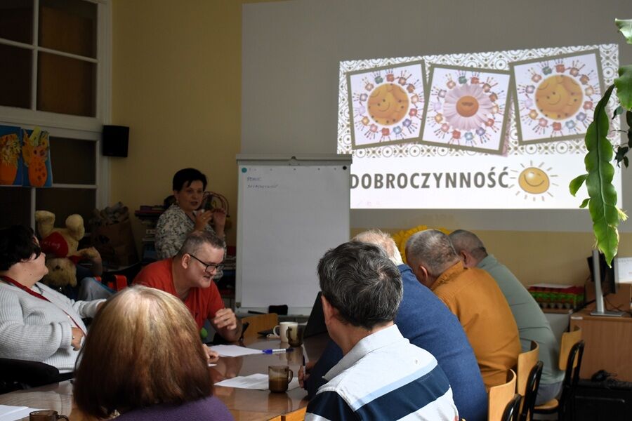 Grupa osób siedzi przy stole podczas zajęć edukacyjnych. Na ścianie wyświetlana jest prezentacja z napisem „Dobroczynność” i rysunkami uśmiechniętego słońca. Prowadząca stoi obok tablicy.