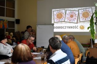 Grupa osób siedzi przy stole podczas zajęć edukacyjnych. Na ścianie wyświetlana jest prezentacja z napisem „Dobroczynność” i rysunkami uśmiechniętego słońca. Prowadząca stoi obok tablicy.