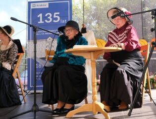 Starsze kobiety w strojach stylizowanych na dawne, eleganckie suknie i kapelusze siedzą przy małym okrągłym stoliku na scenie. Jedna z nich mówi do mikrofonu.