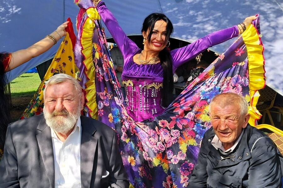 Dwaj starsi mężczyźni siedzą na zewnątrz, obok nich kobieta w barwnej, rozłożystej sukni w kwiaty z podniesionymi szeroko rękawami i trenem.