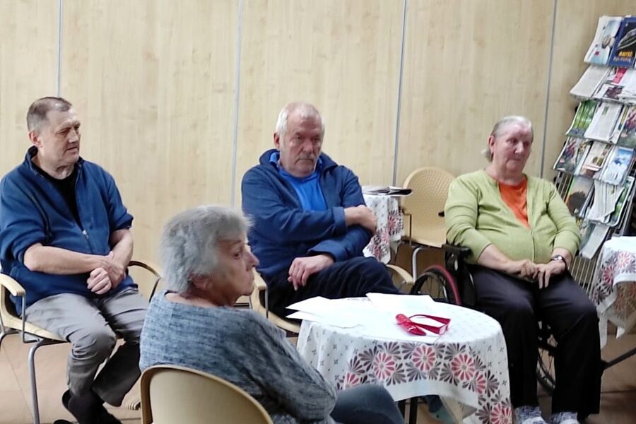Grupa starszych osób siedzi w sali z jasnymi drewnianymi ścianami. Rozmawiają lub słuchają, przy stolikach nakrytych białymi obrusami w kwiatowy wzór. W tle widać stojak z kolorowymi czasopismami.