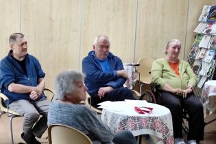 Grupa starszych osób siedzi w sali z jasnymi drewnianymi ścianami. Rozmawiają lub słuchają, przy stolikach nakrytych białymi obrusami w kwiatowy wzór. W tle widać stojak z kolorowymi czasopismami.