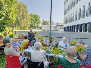 Na zdjęciu Mieszkańcy siedzący przy stołach z leżącymi kwiatami, roślinami do robienia bukietów. 