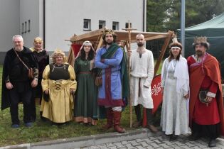 Grupa osób w średniowiecznych strojach – sukniach, pelerynach i koronach – stoi na zewnątrz przed drewnianą konstrukcją namiotu, w tle budynek i drzewa.