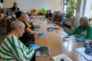 Kilka starszych osób siedzi przy długim stole w jasnej sali i tworzy prace plastyczne, używając czarnych farb na folii aluminiowej, w rękawiczkach ochronnych.

