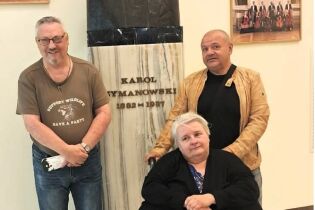 Trzy osoby pozują przy marmurowym pomniku z napisem „Karol Szymanowski 1882–1937” we wnętrzu  filharmonii.

