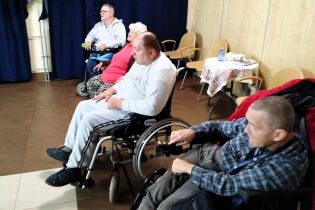 Kilka osób, w tym dwie wózkach inwalidzkich, siedzi w pomieszczeniu z brązową podłogą i granatowymi zasłonami. W tle ustawione są krzesła i stolik z koronkową serwetą oraz butelką wody. 