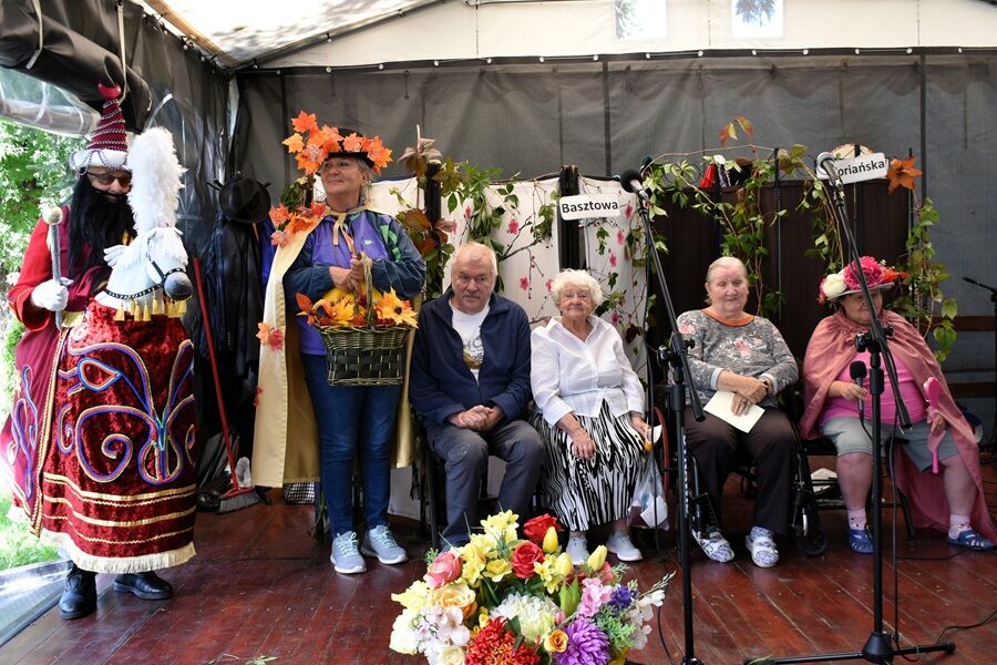 Scena na zadaszonej estradzie udekorowanej roślinami i kwiatami. Kilka osób starszych siedzi w rzędzie, a obok nich stoją dwie osoby w kolorowych kostiumach.