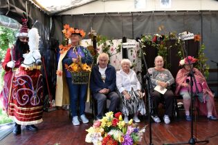 Scena na zadaszonej estradzie udekorowanej roślinami i kwiatami. Kilka osób starszych siedzi w rzędzie, a obok nich stoją dwie osoby w kolorowych kostiumach.