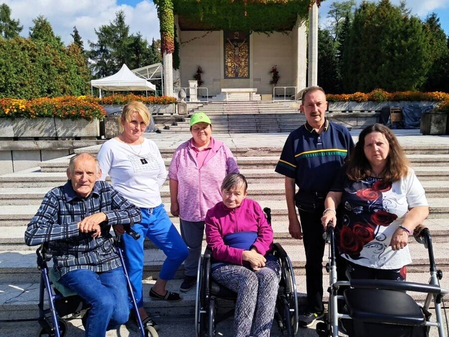 sześć osób stojących i siedzących na schodach przed budynkiem z ołtarzem ozdobionym mozaiką i zielenią. 