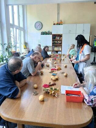 grupa osób siedzących przy długim, drewnianym stole w jasnej sali z dużymi oknami. Na stole rozłożone są różne przedmioty: ziemniaki, kolorowe liście, nasiona, kredki i drobne materiały plastyczne. 
