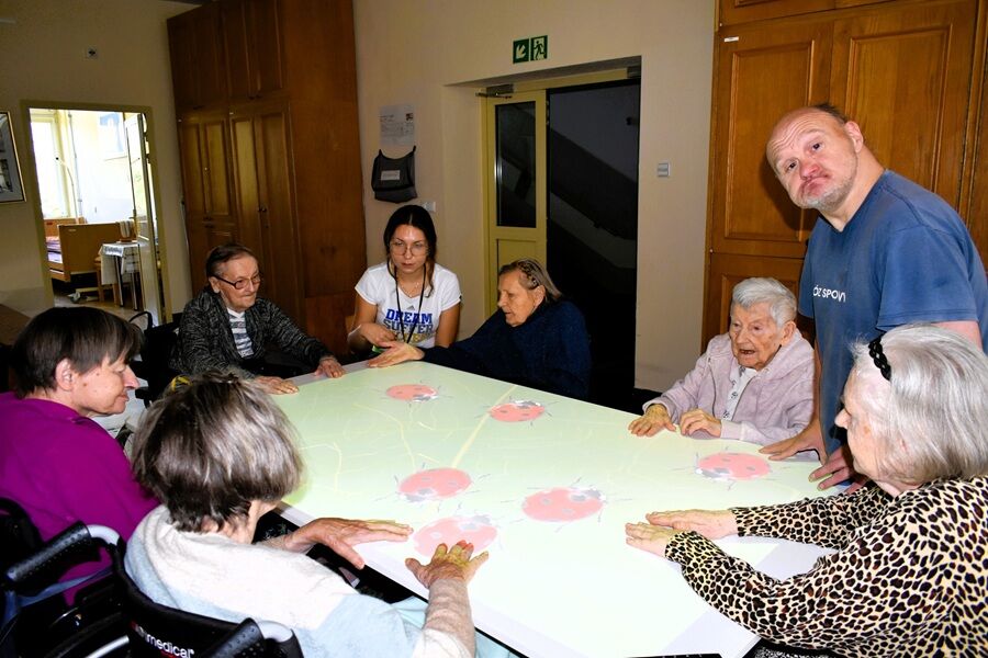 siedem osób siedzących, jedną stojącą wokół dużego, interaktywnego stołu w sali zajęciowej Domu Pomocy Społecznej. Uczestnicy biorą udział w zajęciach terapeutycznych.
