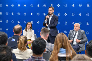 Fotografia ze spotkania Prezydenta Krakowa z cudzoziemcami. W centrum zdjęcia Prezydent Aleksander Miszalski przemawia stojąc i trzymając mikrofon bezprzewodowy. W pierwszym planie głowy uczestników. 