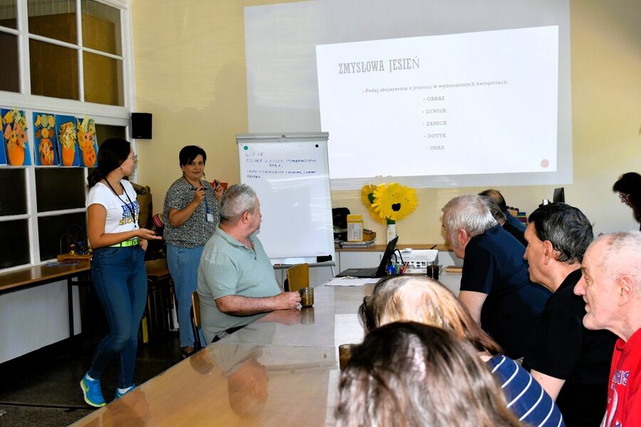 Grupa osób siedzi przy stole podczas zajęć „Zmysłowa jesień”. Na ekranie widać slajd z pytaniami, a prowadzące zapisują pomysły uczestników na tablicy.