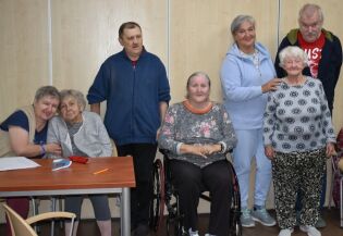 Kilka osób, w tym seniorzy i opiekunowie, pozuje wspólnie do zdjęcia w sali. Jedna z kobiet siedzi na wózku, reszta stoi obok w przyjaznej atmosferze.