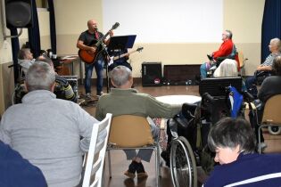 Koncert na żywo: muzyk gra na gitarze i śpiewa, a przed nim siedzi publiczność, w większości seniorzy, niektórzy na wózkach inwalidzkich.