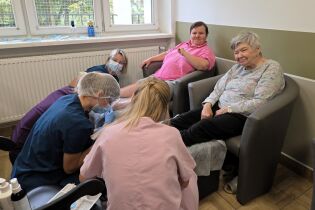 Starsze kobiety siedzą w fotelach, podczas gdy pielęgniarki i podolodzy w maseczkach zajmują się ich stopami, wykonując zabiegi pielęgnacyjne.