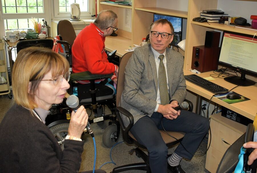 Mężczyzna w garniturze i kobieta z mikrofonem siedzą przy komputerach w pokoju radiowęzła, obok nich mężczyzna na wózku inwalidzkim.