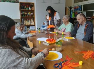 Grupa seniorów siedzi przy stole i wykonuje prace plastyczne z kolorowego papieru pod opieką instruktorki arteterapii.