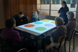 Grupa seniorów siedzi wokół stołu multimedialnego z kolorowymi kółkami wyświetlanymi na blacie. Uczestnicy z zainteresowaniem biorą udział w zajęciach sensorycznych, a obok nich stoi prowadząca.