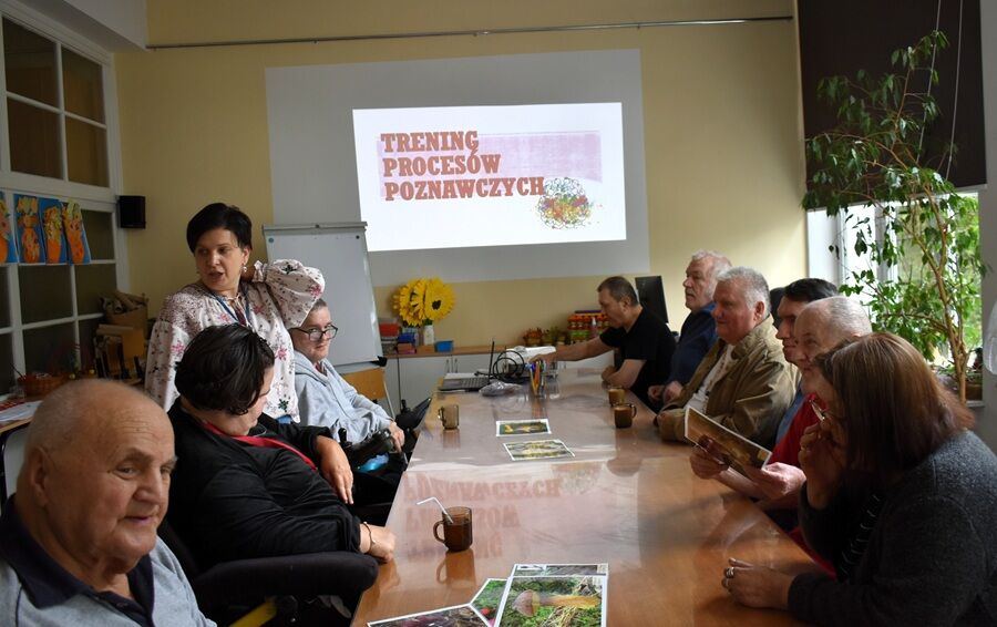 Seniorzy siedzą przy stole w świetlicy, przed nimi materiały dydaktyczne i napoje. Na ścianie wyświetlony jest napis „Trening procesów poznawczych”. Zajęcia prowadzi terapeutka.