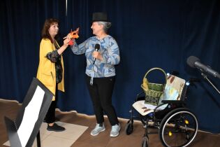 Dwie kobiety biorą udział w występie artystycznym. Jedna z nich trzyma mikrofon i koszyk, druga wręcza jej jesienne liście. W tle ciemna kotara, obok stoi wózek inwalidzki.