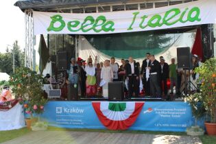 Grupa osób na scenie plenerowej udekorowanej włoskimi barwami. W tle widoczne są dekoracje, a na scenie występują uczestnicy w różnych strojach. Przed sceną widoczne są rośliny w donicach.