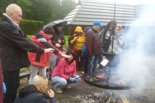 Grupa osób zgromadzona wokół dużego, okrągłego grilla nad ogniskiem. Na ruszcie pieką się kiełbaski, a dwaj mężczyźni grzeją zmarznięte dłonie przy ogniu. 