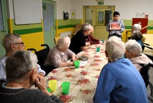 Grupa starszych osób siedzi przy długim stole w sali zajęciowej. Instruktorka stoi na końcu stołu i pokazuje obrazek, a uczestnicy uważnie go oglądają. Na stole leży obrus w kwiaty.