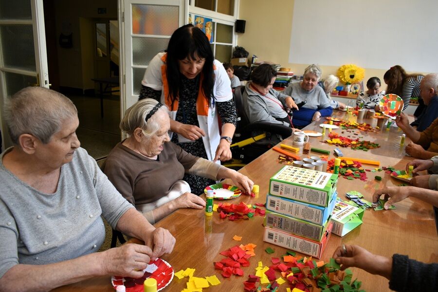 Starsze osoby siedzą przy stole i wykonują prace plastyczne z papieru kolorowego. Opiekunka pomaga jednej z uczestniczek. Na stole leżą kolorowe kawałki papieru, kleje i pudełka po produktach.