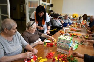 Starsze osoby siedzą przy stole i wykonują prace plastyczne z papieru kolorowego. Opiekunka pomaga jednej z uczestniczek. Na stole leżą kolorowe kawałki papieru, kleje i pudełka po produktach.