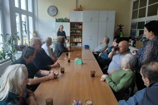 Grupa seniorów siedzi przy długim stole i słucha kobiety prowadzącej zajęcia. W tle widać szafki, półki z materiałami plastycznymi i duże okna wpuszczające światło dzienne.