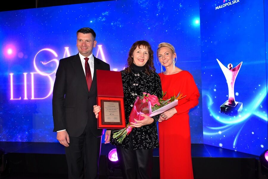 Trzy osoby stoją na scenie podczas uroczystej gali. Kobieta w środku trzyma kwiaty i tabliczkę z wyróżnieniem, a po bokach stoją elegancko ubrany mężczyzna i kobieta w czerwonej sukni. 
