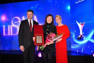 Trzy osoby stoją na scenie podczas uroczystej gali. Kobieta w środku trzyma kwiaty i tabliczkę z wyróżnieniem, a po bokach stoją elegancko ubrany mężczyzna i kobieta w czerwonej sukni. 