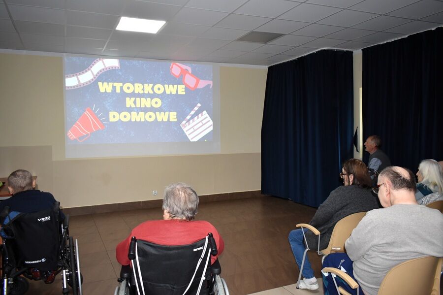Kilka osób, w tym osoby na wózkach inwalidzkich, ogląda film wyświetlany na dużym ekranie w sali. Na ekranie widać napis „Wtorkowe Kino Domowe” oraz ilustracje klapsa filmowego i okularów 3D.