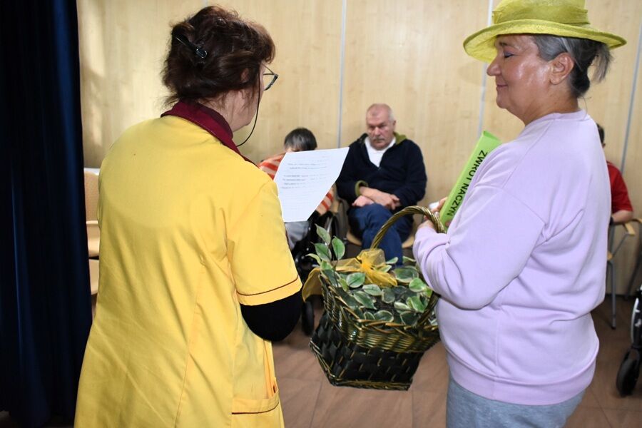 Dwie kobiety stoją naprzeciw siebie w sali — jedna w żółtym fartuchu czyta z kartki, druga w jasnozielonym kapeluszu trzyma koszyk ozdobiony liśćmi. W tle siedzi kilka osób obserwujących rozmowę.