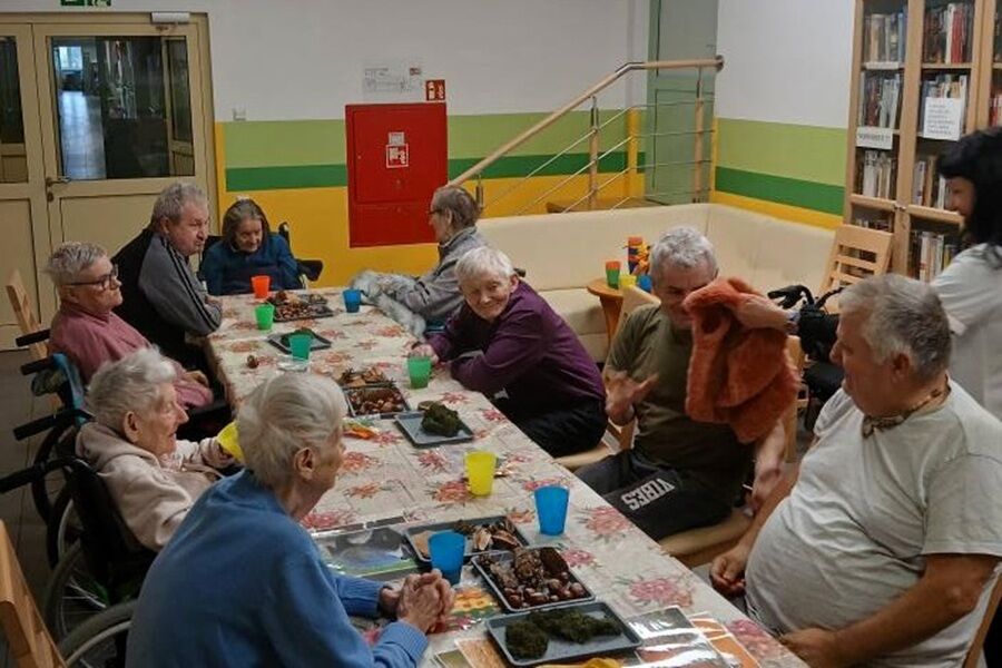 Grupa starszych osób siedzi przy długim stole przykrytym obrusem w kwiaty. Na stole stoją kolorowe kubki i tace z różnymi materiałami, które wyglądają jak elementy do zajęć sensorycznych.