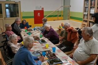 Grupa starszych osób siedzi przy długim stole przykrytym obrusem w kwiaty. Na stole stoją kolorowe kubki i tace z różnymi materiałami, które wyglądają jak elementy do zajęć sensorycznych.