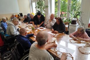 Kilkunastoosobowa grupa seniorów bierze udział w zajęciach arteterapii przy dużym stole. Uczestnicy wykonują prace plastyczne z papieru i naturalnych materiałów, wspomagani przez opiekunkę. 