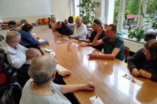 Seniorzy siedzą wokół dużego stołu i grają w grę towarzyską przypominającą lotto lub bingo. Na stole leżą karty z liczbami i kolorowe żetony. Uczestnicy są skupieni i zaangażowani.