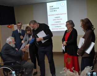 Kilka osób stoi na scenie podczas uroczystości wręczenia nagród literackich. Mężczyzna na wózku odbiera dyplom od prowadzących. W tle wyświetlony jest slajd z tytułem konkursu „Dom Pięknego Słowa”. 