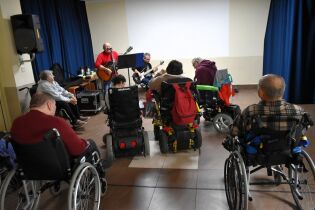 Grupa osób na wózkach uczestniczy w koncercie lub warsztatach muzycznych. Dwóch muzyków gra na gitarach i śpiewa, a uczestnicy słuchają ich z uwagą.

