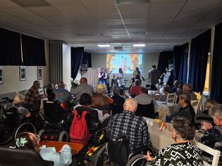 Grupa osób starszych, głównie na wózkach inwalidzkich, siedzi w sali z ciemnymi zasłonami, oglądając występ muzyczny na żywo. Na scenie stoi kilku wykonawców z instrumentami.