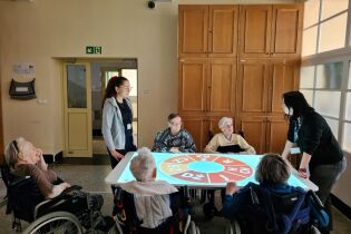 Kilka osób starszych siedzi wokół stołu interaktywnego w jasnym pomieszczeniu. Na stole wyświetlany jest kolorowy ekran z literami i symbolami. 