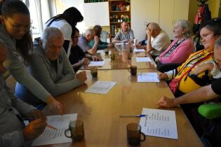 Seniorzy siedzą przy dużym stole, wykonując ćwiczenia poznawcze na kartkach papieru. Dwie opiekunki pomagają im, wskazując odpowiedzi. Na stole stoją kubki, a w tle widać szafki i półki.