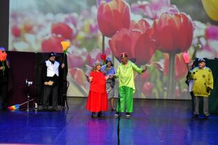 Grupa aktorów występuje na scenie w kolorowych kostiumach przedstawiających owady i kwiaty. W tle widać duży ekran z obrazem tulipanów.