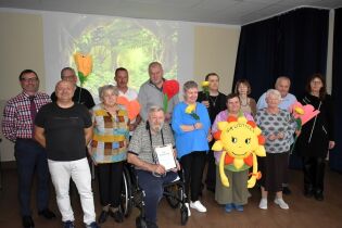 Grupa uczestników pozuje razem do zdjęcia, trzymając kolorowe kwiaty i maskotkę Gaudynkę w kształcie uśmiechniętego słońca. Na pierwszym planie widoczny dyplom za Nagrodę Publiczności.