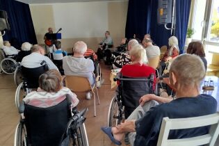 Grupa seniorów, głównie na wózkach inwalidzkich, siedzi w sali i słucha mężczyzny grającego na gitarze. Muzyk stoi przy mikrofonie na tle granatowej zasłony. 