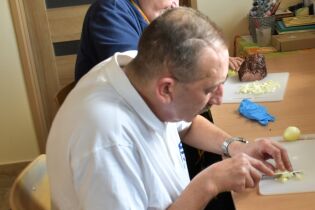 Dwóch mężczyzn siedzi przy stole i przygotowuje posiłek. Jeden z nich kroi cebulę na desce do krojenia, a w tle widać kawałek mięsa oraz kuchenne akcesoria.

